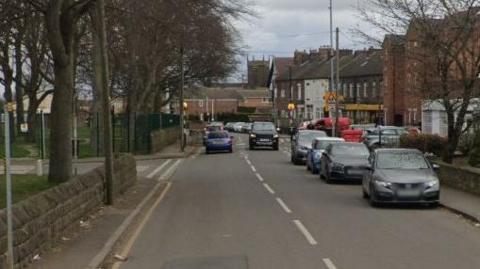 A road in Wakefield, with a pedestrian crossing and and row of shops in view. A church spire can be seen in the distance. 