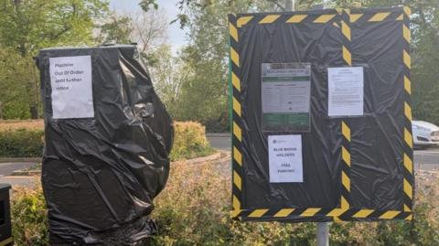 A parking machine covered in black tape, with a sign on it, next to a sign also covered in black plastic, with a number of notices on it. A car is behind the sign, by buses and trees. 