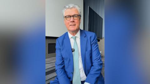 Reform UK Scotland leader Malcolm Offord smiles at the camera as he sits in front of an office background. He has silver hair, blue eyes and is wearing glasses, a light blue tie, a white shirt and a blue suit.