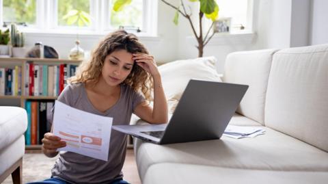 A woman has her hand pressed against her head as she sits on the floor with her laptop on the sofa looking at her energy bill on a piece of A4 paper