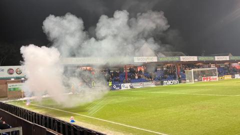 Smoke rises from the pyrotechnic as it burns near a corner flag on Ross County's pitch in Dingwall. The ground's Jail End stand is in the background.