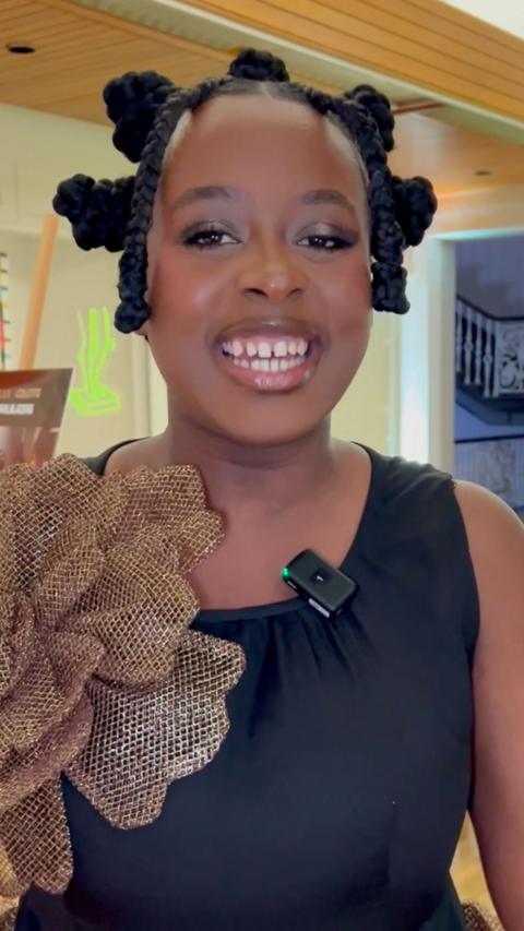 A smiling girl, with a one shoulder dress and braided hair which sits in 7 buns on her head.