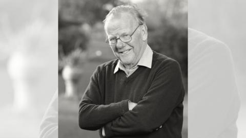 A black and white picture of a man with glasses and thinning hair. He is smiling and crossing his arms. He is wearing a dark jumper over a shirt.