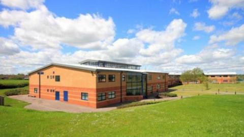 A modern two-storey L-shaped school building in the middle of a field. It has orangey-red brickwork, which appears striped on the ground floor, and there are blue doors. The sky above is blue with some white clouds.