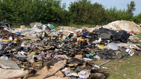 Large piles of dumped and burnt waste spread across open land in Northfleet, including household rubbish, carpets, timber, plastic containers, blackened debris and construction materials, with trees and fencing in the background.