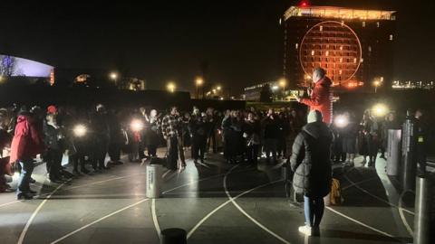 A group of people stood in the dark, lit by mobile phone camera lighting, with Milton Keynes Hotel Le Tour lit up in the background.