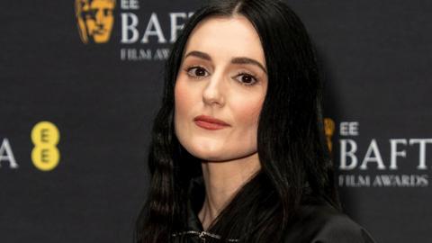 Actress Posy Sterling, with long black hair and red lipstick, looks at the camera in front of a wall bearing EE Bafta Awards branding