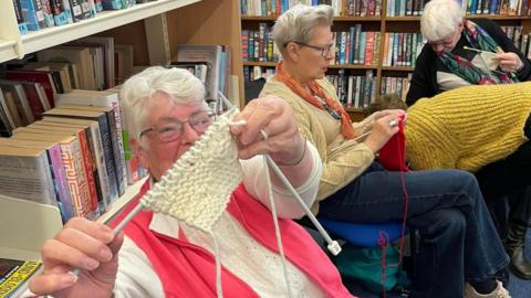 A group of three ladies are sitting on chairs in the library knitting, one of them has pointed her knitting needles and a small piece of knitted fabric and the camera