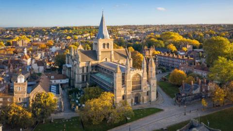An aerial shot of Rochester Cathedral. There are dozens of green trees to either side, and a number of other properties. 