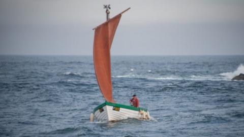 A white boat with orange sail upon the sea with a man in an orange jacket aboard. There are waves crashing against rocks to the right of the photograph