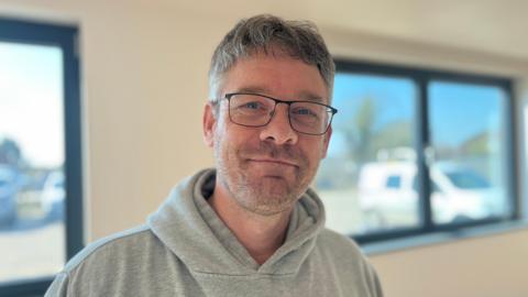 A man with grey hair wears a grey hooded top and black-framed glasses as he smiles for the camera with the white walls and grey-framed windows of a village hall seen behind him in soft focus.