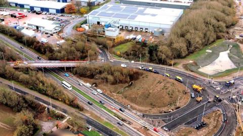 An aerial view of a construction site for a new junction. A dual carriageway can be seen running alongside the bottom of the photo with a partially-constructed road shooting off to one side of it. There are some industrial buildings in the background.