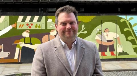 Seb Slater is dressed in a light grey coat, with a white shirt. He has light brown hair, stubbly facial hair and is smiling. In the background, there is a mosaic painted on boards. It shoes people collecting food, including one with a green shirt, navy blue apron and a roast chicken on a plate.
