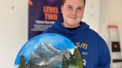 A young person in a blue hoody holds a circular canvas with a painting of mountains, blue sky and trees,