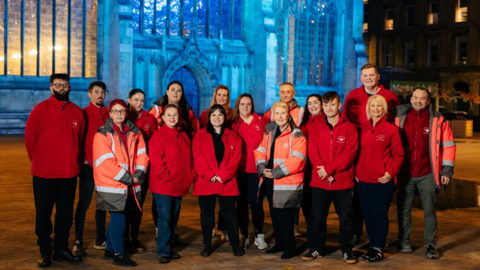 Group shot of volunteers from City of Hull Street Angels standing in front of a large stone church lit up in blue. People in the group are wearing red fleeces with the street angels logo - which has wings to either side - visible on one side.