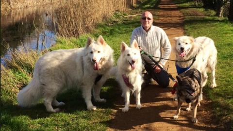 Matthew Young kneeling down holding four dog leads. He is wearing a white knitted jumper and dark jeans. Three dogs are white furred and one smaller dark haired dog.