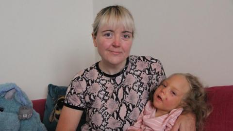 A woman in a pink alligator skin print t-shirt, with bleach blonde hair smiles at the camera, who has honey blonde hair, and looks at the camera. They are sitting on a red sofa with a blue stuffed toy next to them and a white wall behind. 