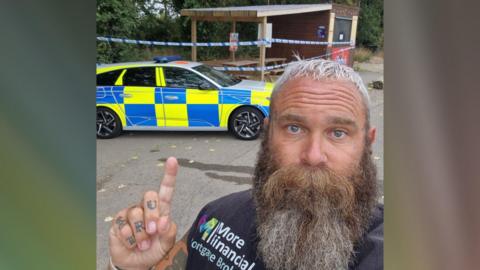 Adam wearing a black T-shirt points his finger to the crime scene behind him - with a police car and blue police tape around a wooden food shed.