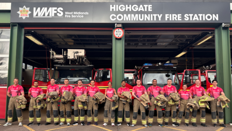 The male and female firefighters stand in a line at the entrance to their station with fire engines behind them. They are wearing pink T-shirts and their brown/black uniform trousers and are holding the top jackets.