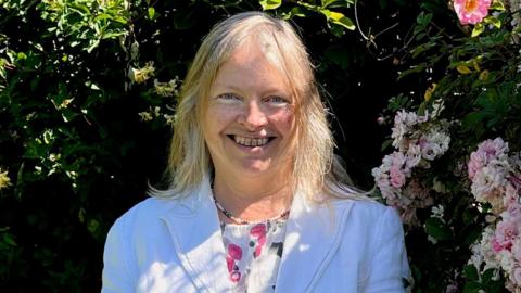 Annabel Yates stood outside next a rose bush. She is looking straight at the camera with a wide smile on her face. 