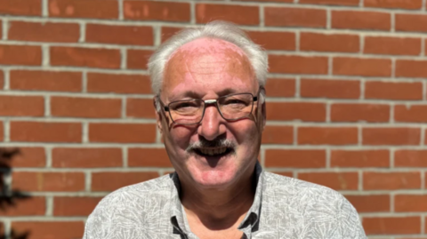 A man with glasses and grey hair smiling into the camera while standing in front of a brick wall.