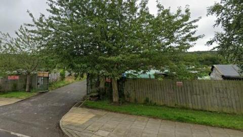 A wooden fence which lines a path and road. A large tree is at the entrance of an open wooden gate. An allotment can be seen behind the fence.