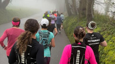 A group of people seen from behind, running on a road in Jersey. They are wearing colourful tops and running vests.