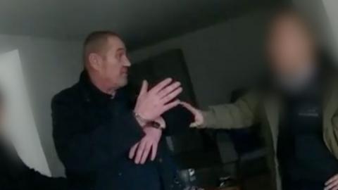 A man in handcuffs is being held by another person with a blurred face in a dimly lit room.