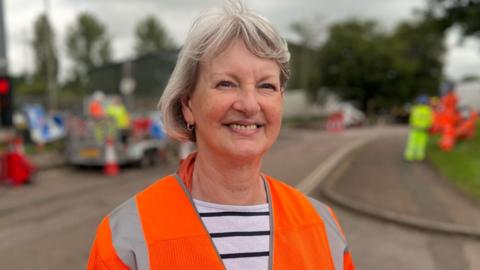 Liz Leffman has medium length grey hair and is smiling. She is wearing an orange hi-vis over a white and blue striped t-shirt. Behind her, contractors are fixing potholes.