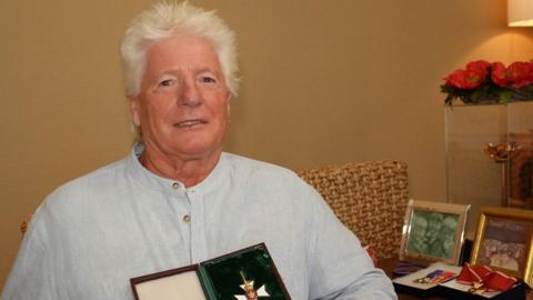 Bruno Peek who has white hair and is wearing a pale blue striped collarless shirt. He is sitting down and is holding up a case in which an order can be seen. There is a desk to his left with photos and more honours