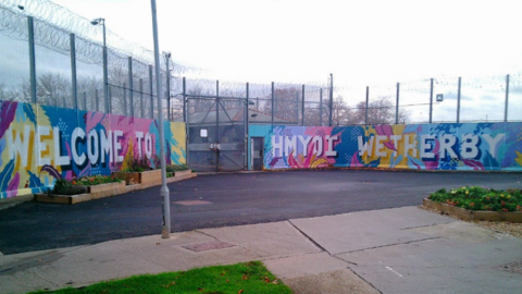 The image shows the exterior of Wetherby Young Offender Institution with tall metal fencing topped with rolls of barbed or razor wire. A painted mural covers the perimeter wall, featuring colourful patterns and large white lettering that reads: "Welcome to HMYOI Wetherby". The ground is paved, with small planted flower beds in front of the mural.