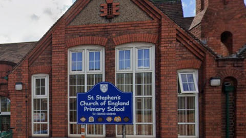 The school is a red brick building close to a road. It has large windows and a blue sign outside displaying the name of the school.
