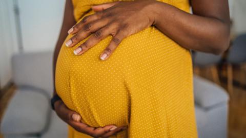 Close-up of pregnant black woman wearing a yellow dress with small white dots. The picture does not show the woman's face. She has both hands resting on her pregnant belly and she is standing in a room with a grey sofa behind her and some grey chairs and a table.