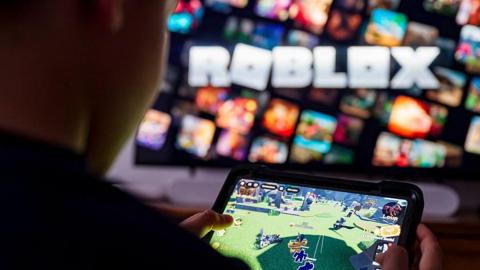 An over the shoulder view of a child holding a tablet playing a game on Roblox. In the background is a blurred screen with the Roblox logo on it.