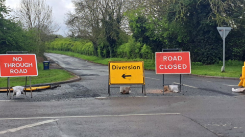 Three road signs warning of a closure and diversion block Barnhurst Road. The road is joined to a larger road. Trees line either side of Barnhurst Road which has no road markings on.