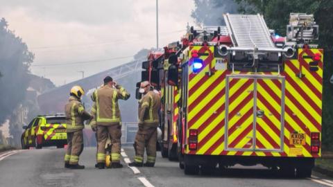 A row of fire engines and four firefighters in the foreground, on a road. In the distance is a fire car.