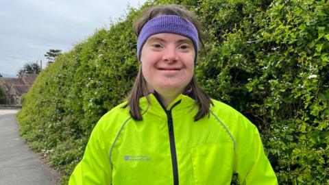 Serena has brown hair and a purple sweatband on her head. She is wearing a bright yellow windbreaker and is smiling. She is standing in front of a green bush on a path.