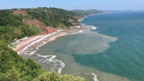 Oddicombe Beach in Torquay - blue sky and sea. Red rockface and green, vegetation covered cliffs
