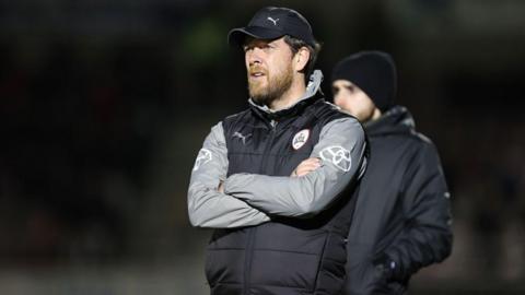 Manager Darrell Clarke is standing with his arms crossed on a pitch. He is wearing a black gilet with grey sleeves visible underneath and a black cap.