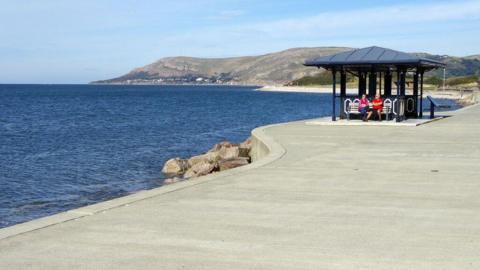 Two people sitting in a hut on a concrete promenade in front of water with hills in the background