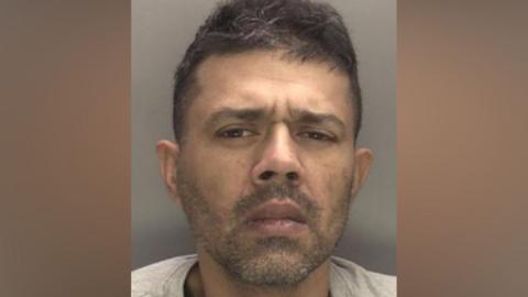 A custody image of Harinderpal Athwal, a man with a short black beard and moustache and short hair is looking at the camera with a slight frown on his face