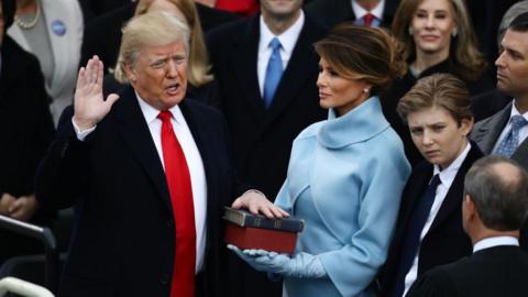 Trump wears a dark suit and red tie. He has one hand resting on a Bible and holds the other up as he recites the oath of office. Melania Trump is dressed in light blue and holds the Bible. They are surrounded by a crowd of people.