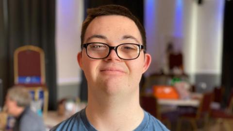 Dylan Smith smiling at the camera in a head and shoulders image. He has dark short hair, black-framed glasses and a blue T-shirt. 