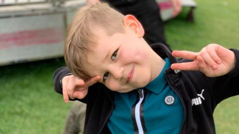 A little boy, who has his head to one side and is smiling. He is wearing a teal top and black hoodie. He is standing in a field and holding both his arms up near his face so his elbows are bent and he is making the peace sign with both of his hands. He has short, light brown hair. 