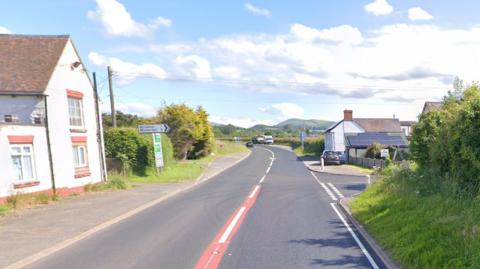 A road with a left turning bend, with a white house on the left hand side and a junction on the right hand side, as well as a white house. Ahead, there are cars parked in a layby