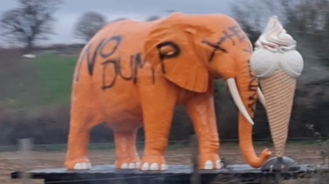 A giant ice cream cone statue next to a large orange elephant statue stood in a field on a Devon farm. The elephant statue has been vandalised with black spray paint with the words "no dump".