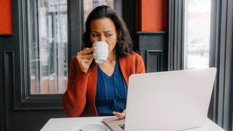 A woman is sipping from a white mug while working on her laptop at a cafe. She appears to be focused, surrounded by a warm interior with red and black accents.