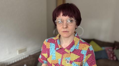 A young woman with short red-brown hair looks at the camera. Behind her is a sofa strewn with cushions. She is wearing large round glasses, even larger hooped earrings and has her left nostril pierced. She is wearing a patterned top with patches of yellow, blue and dark pink