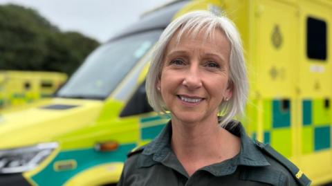 Jackie is stood outside with a yellow ambulance behind her at the ambulance station in Pembroke Dock. She is wearing the dark green ambulance service uniform. She has silvery-white hair in a bob and is smiling at the camera. It is a head and shoulders shot of Jackie.