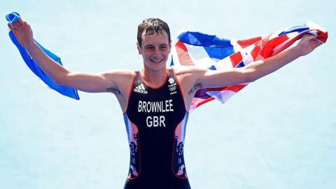 Alistair Brownlee celebrating his gold medal win at the Rio De Janeiro Olympic games in 2016. He is wearing a black Team GB running vest with BROWNLEE GBR written on the chest and black shorts. His arms are outstretched and he is holding the Union Jack flag behind his back, arms and shoulders.